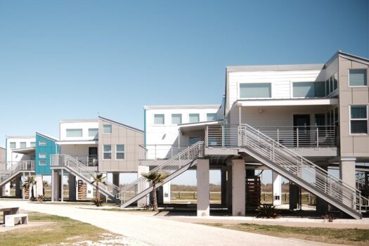 Modern coastal houses raised on stilts, with gray and blue exteriors, large windows, and multiple staircases leading up to entrances. The area is sunny with a clear sky and some small palm trees in front.