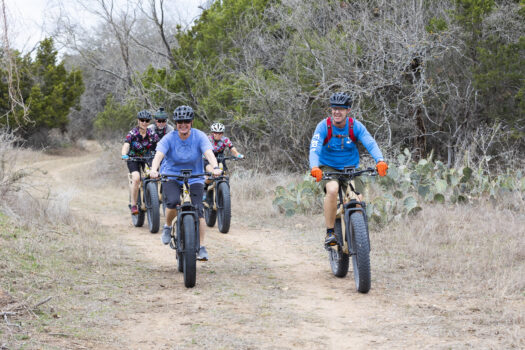 Four cyclists ride fat-tire bikes on a dirt trail through a dry, wooded area. Two are in front smiling, with two more following behind. Leafless trees and cacti are visible along the trail.