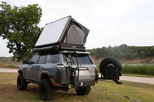 A gray SUV with a rooftop tent set up is parked on grass near a river. The vehicle has off-road tires, a spare tire, and outdoor camping gear attached to the back. Trees and greenery are in the background.